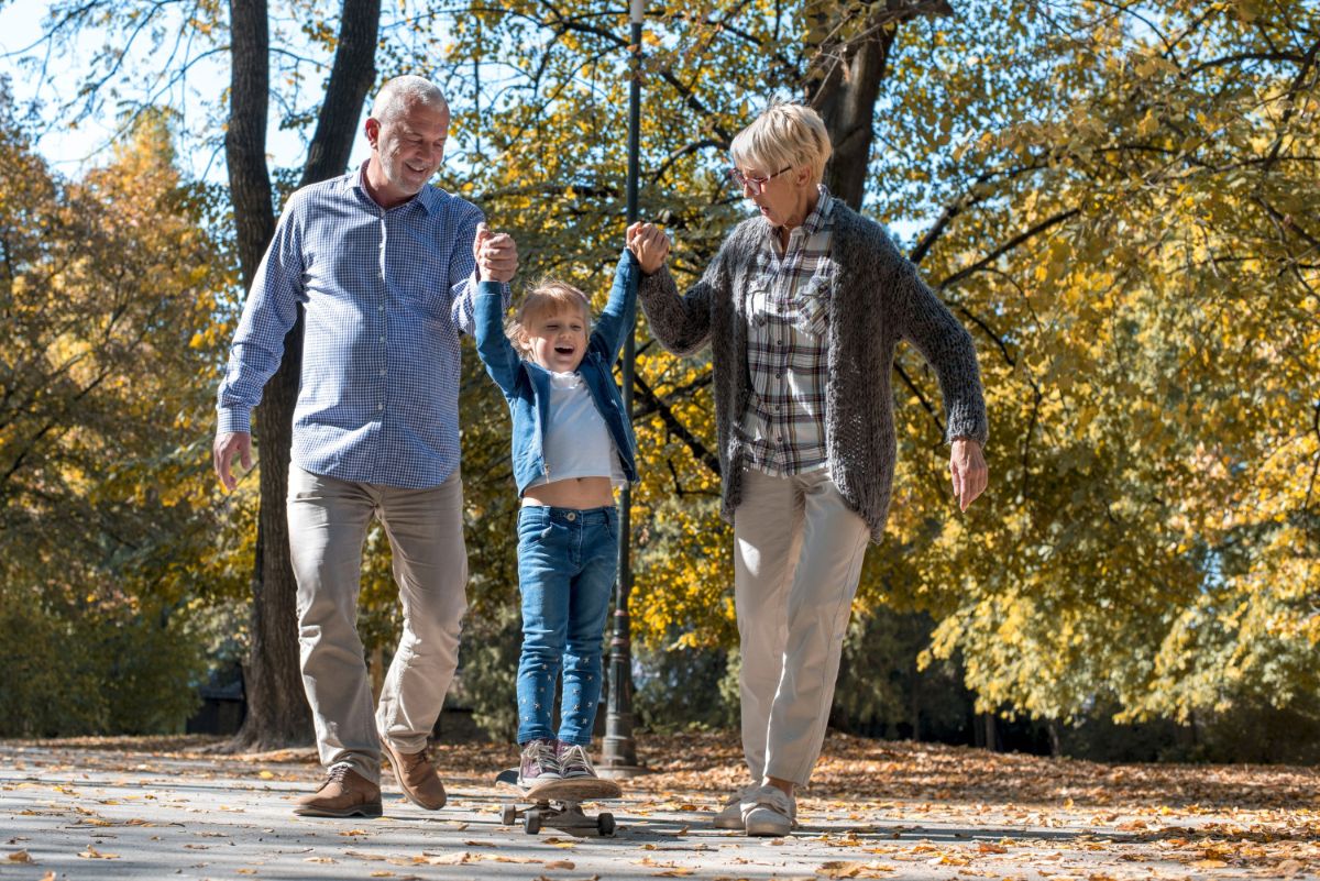 smiling grandparents holding granddaughter s hands while she is riding skateboard autumn park